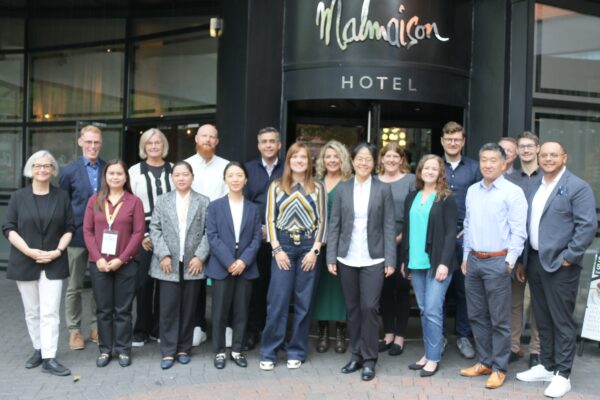 A diverse group of 18 people pose and smile for a photo outside the entrance of Malmaison Hotel, standing on a brick walkway in front of glass doors.